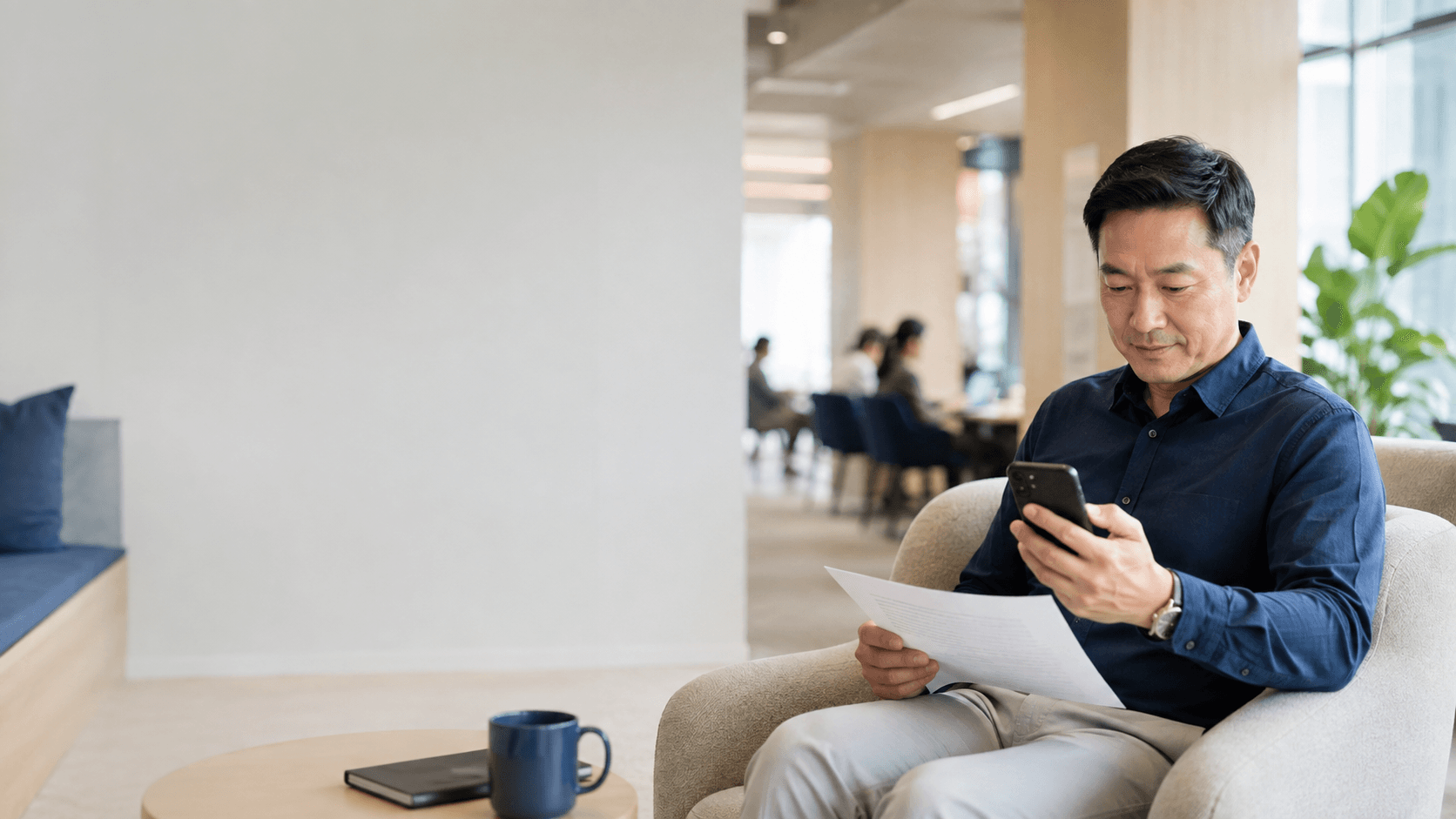 Asian man using a smartphone while holding documents in a bright, modern office lounge.