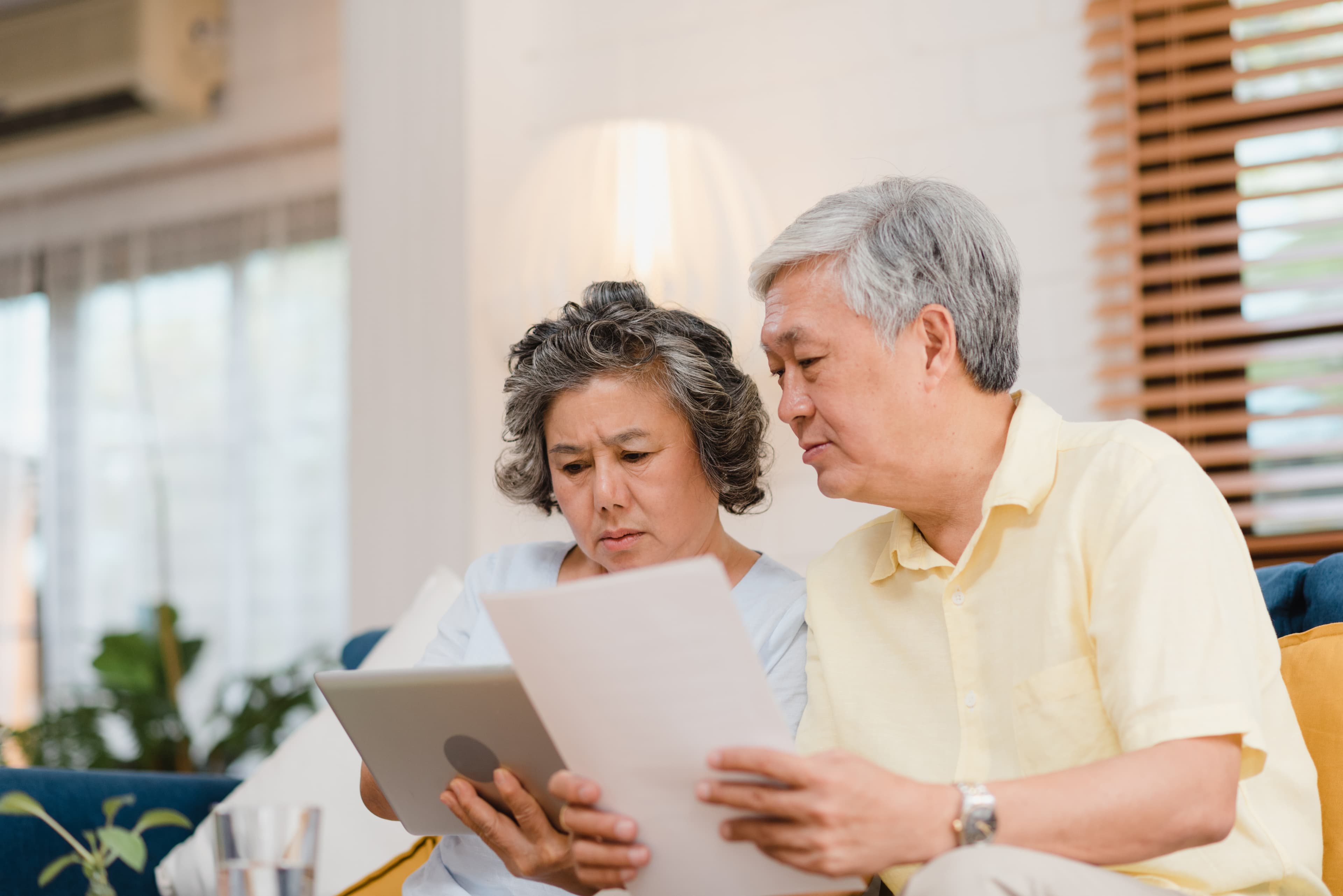 Concerned senior Asian couple reviewing a document and tablet while sitting on a living room sofa.